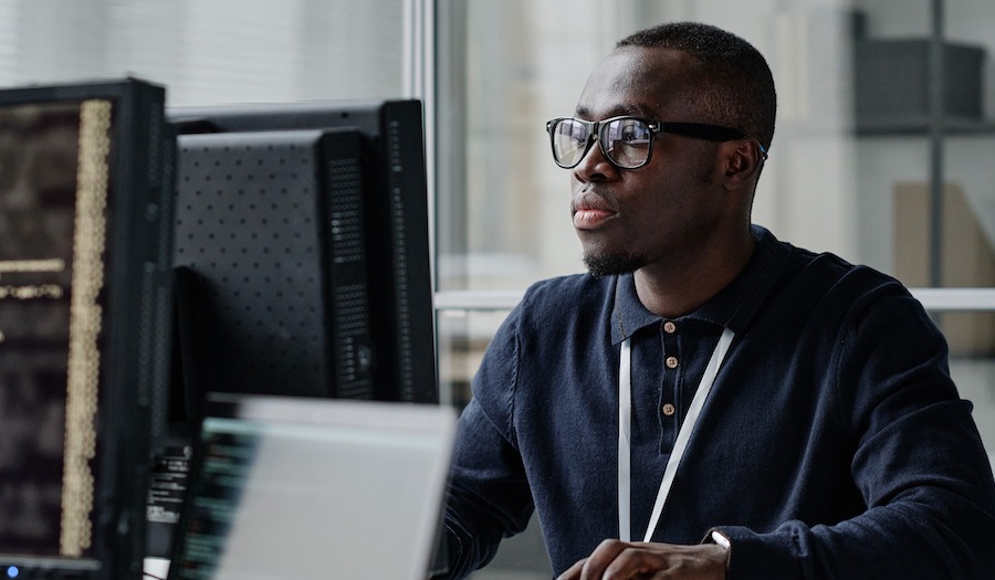 African American young developer in eyeglasses concentrating on his online work on computer sitting at workplace
