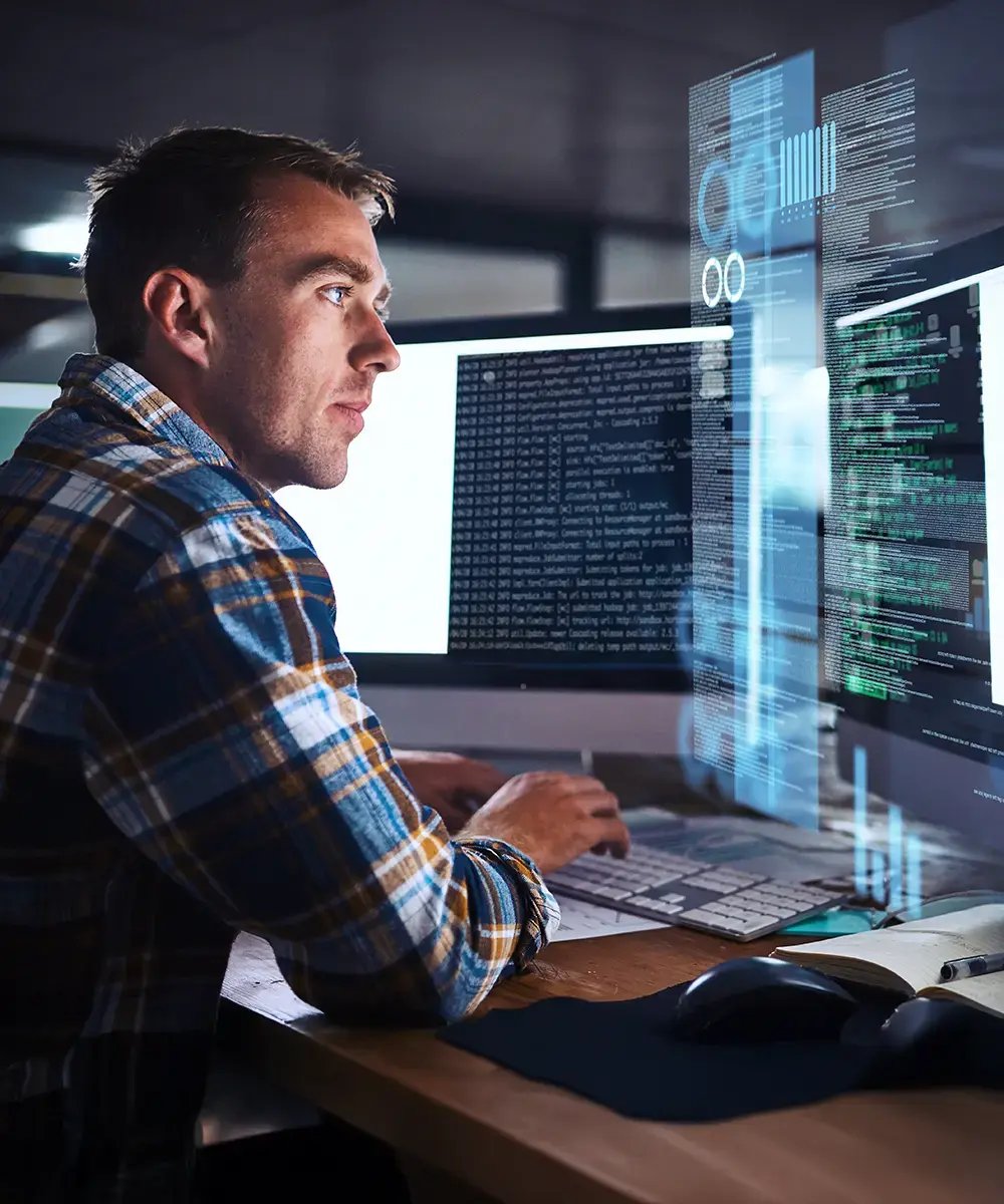 A man in a plaid shirt intensely focuses on a computer screen displaying code and data.