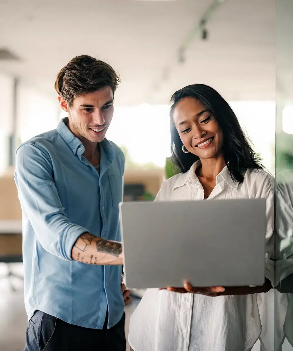 A man and a woman in a bright office smile as they look at a laptop.