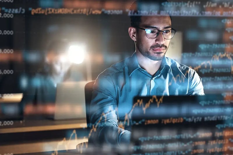 A man in glasses focuses intently on a laptop, surrounded by overlaying digital graphs and numbers.