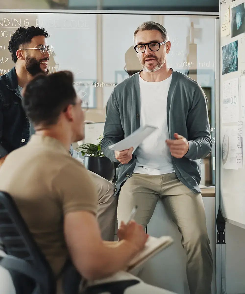 A man is leading a casual meeting in an office, holding papers and speaking to two colleagues.