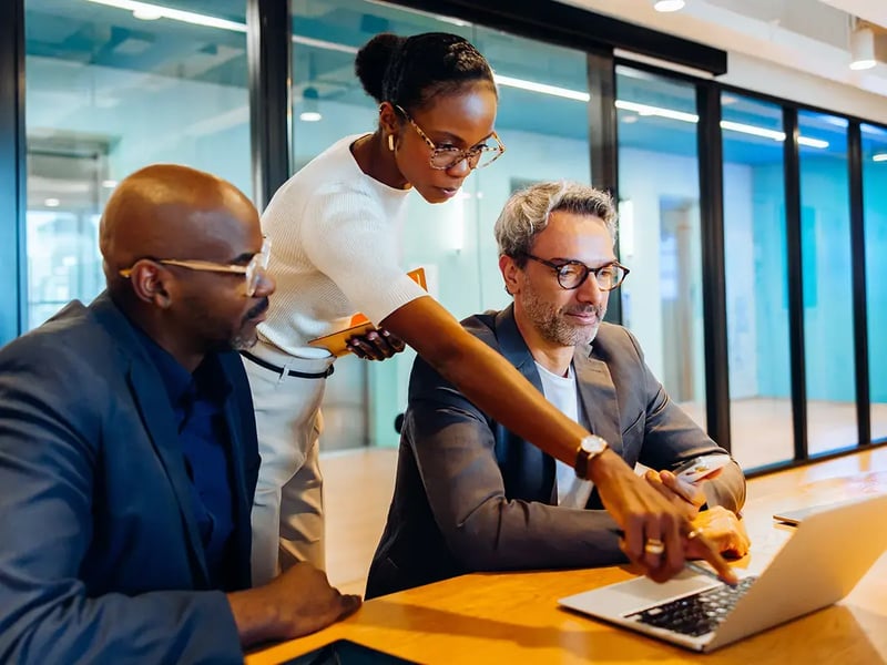 A woman assists two men with a laptop in a modern office, suggesting teamwork