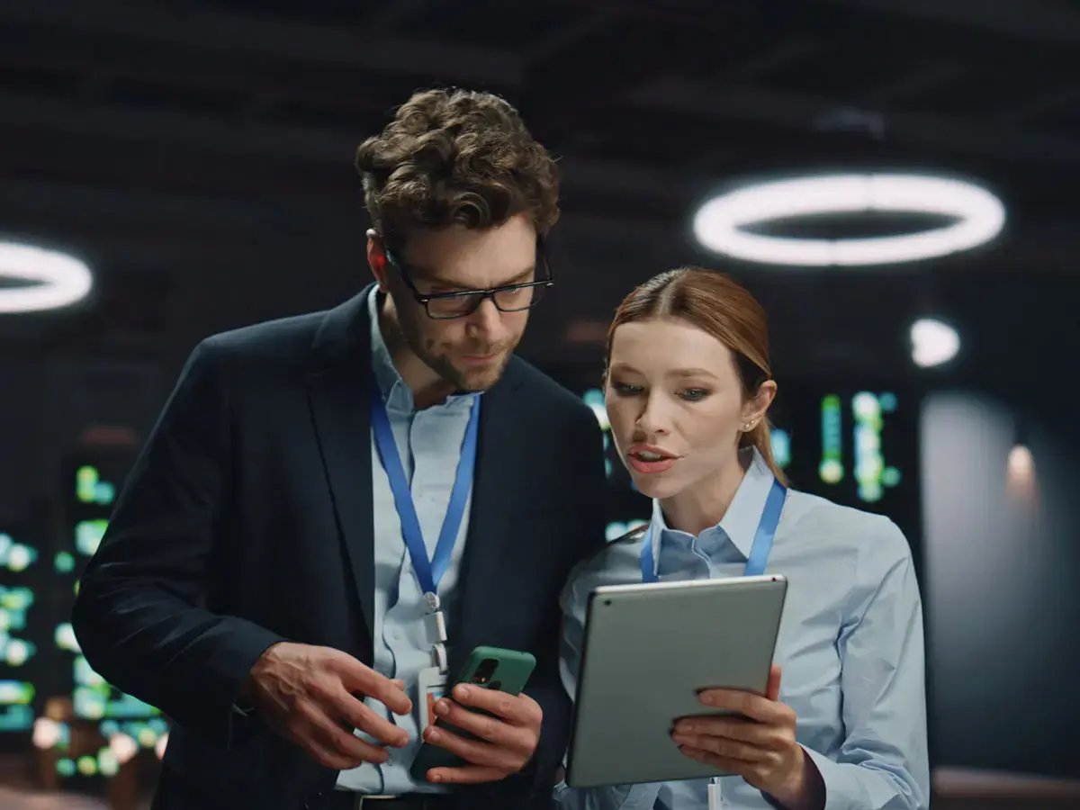 Two IT professionals in an office with digital screens focus intently on a tablet, wearing ID lanyards.