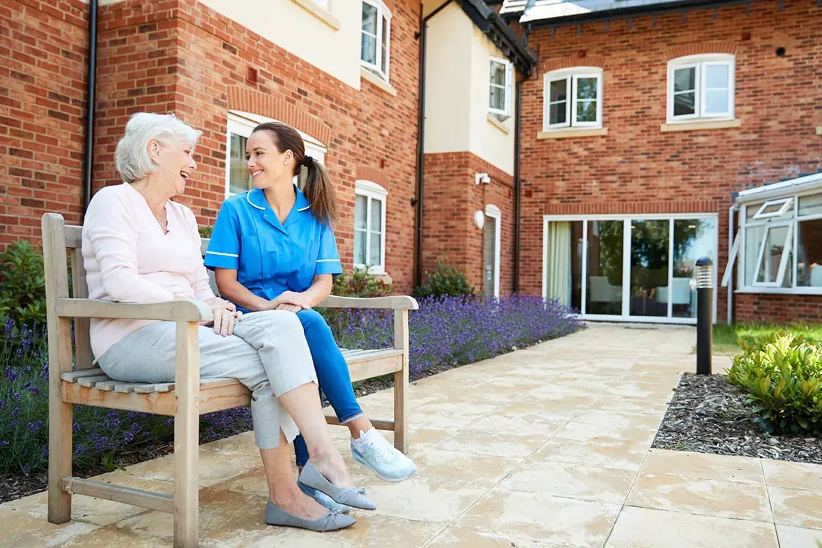 A senior woman sits on a bench alongside a nurse.