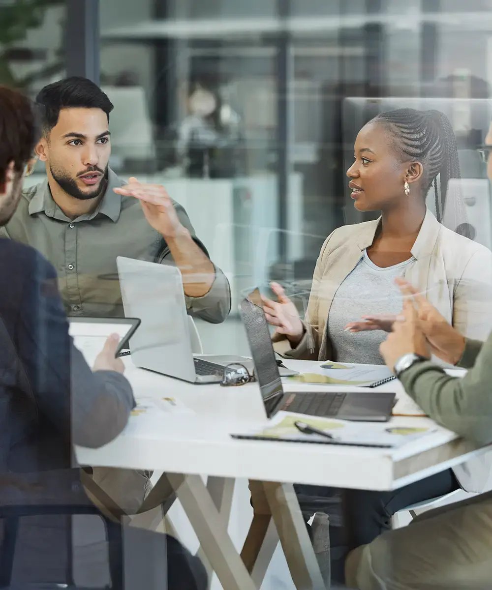 A diverse team engaged in a meeting around a white table, with laptops open.
