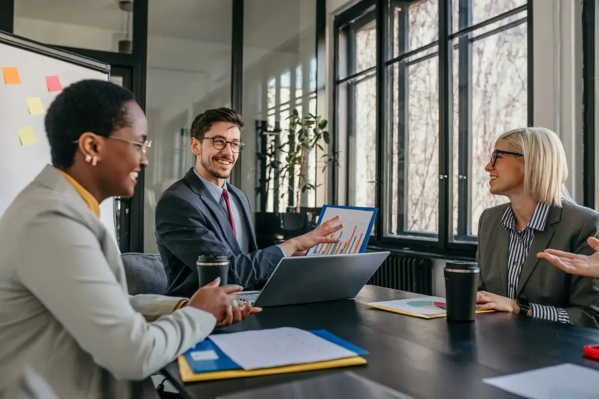 Three colleagues smiling and discussing charts in a modern office, conveying a collaborative and positive atmosphere.