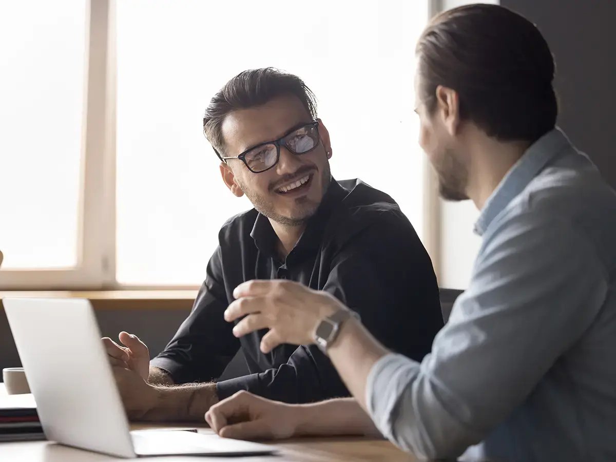 Two men are happily conversing at a desk with a laptop.