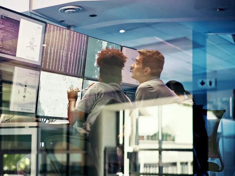 Two people are attentively looking at multiple computer monitors displaying data and graphs in a modern office