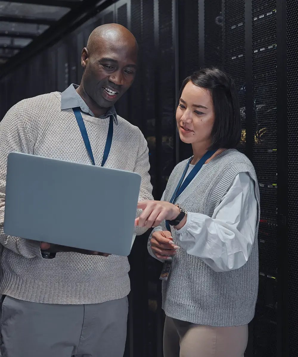 Two people in a server room discuss data on a laptop, both wearing ID lanyards and sweaters.