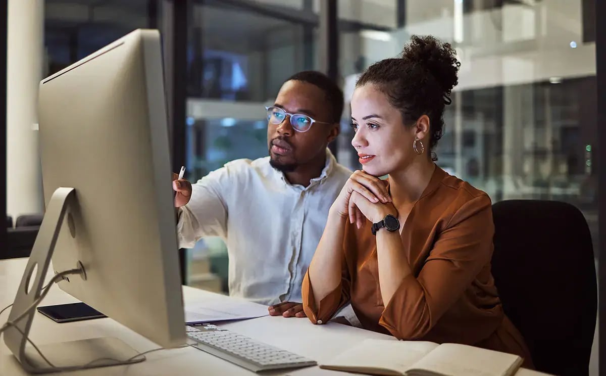Two colleagues collaborate in an office at night, focused on a large computer screen.