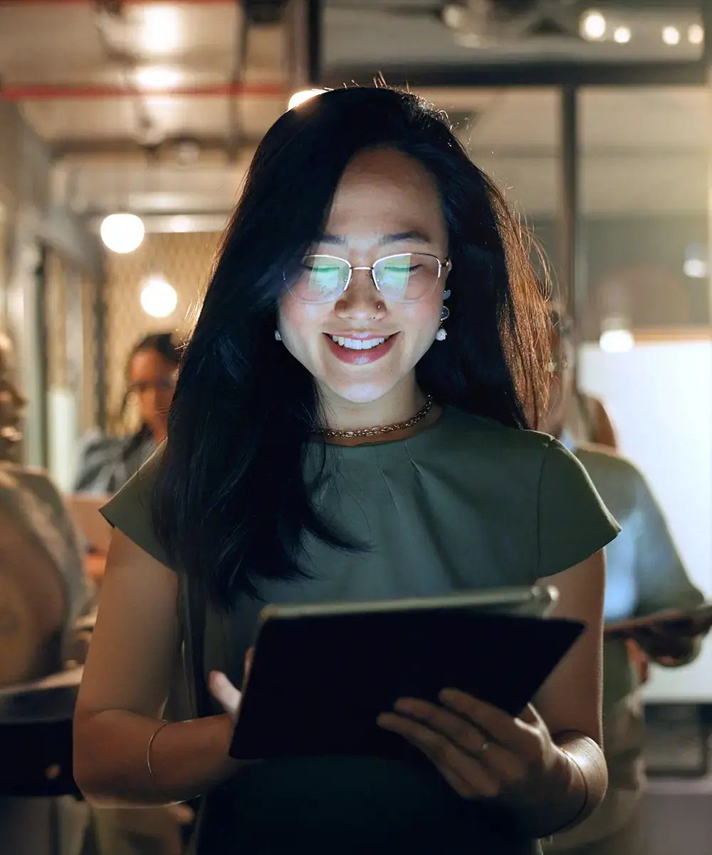Smiling woman in glasses reads a tablet in dimly lit office, surrounded by colleagues.