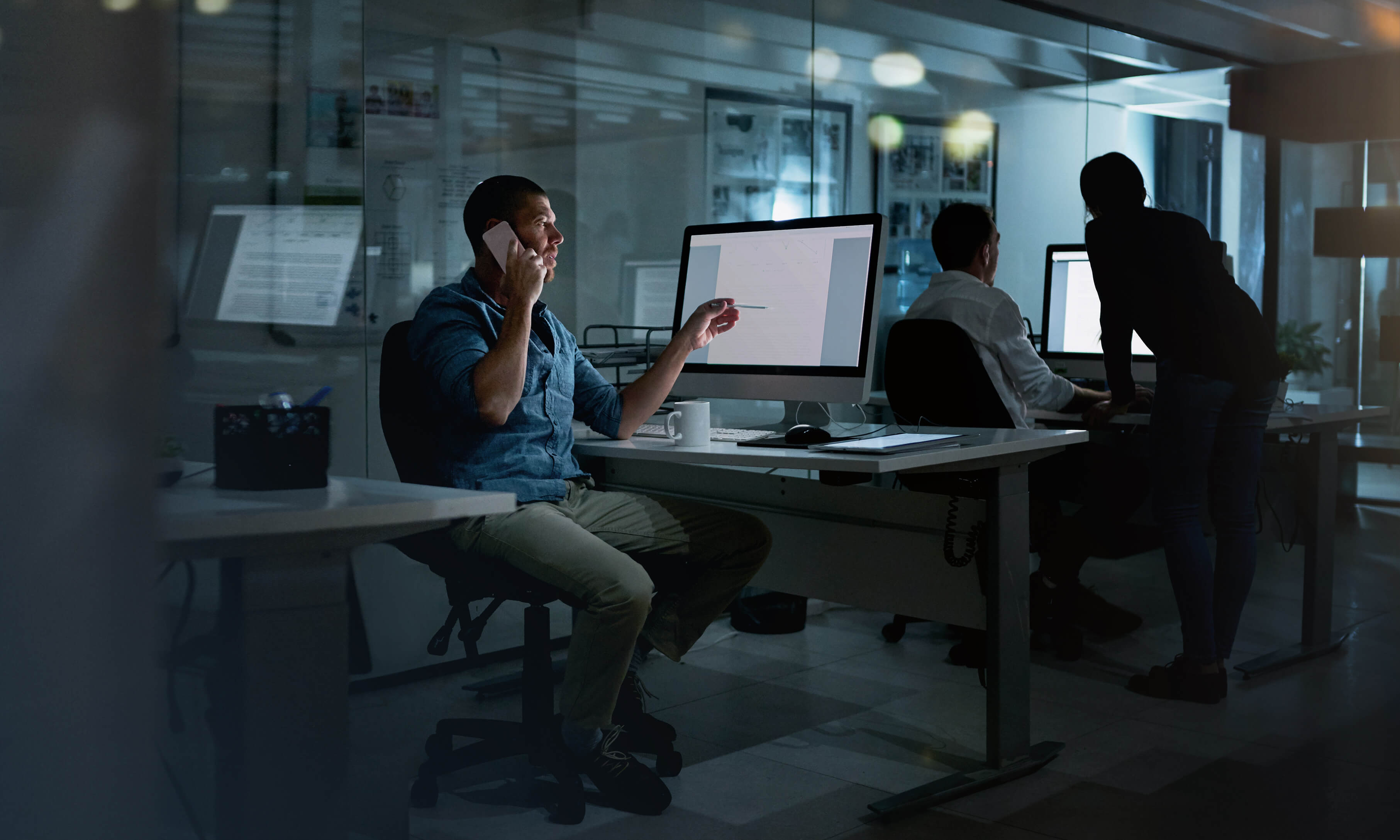 A man talking on the phone while looking at his computer and sitting on his desk at a dark office