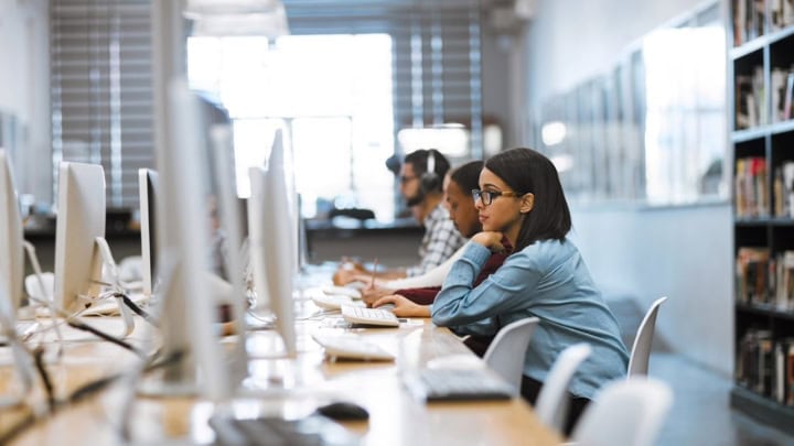 An eye-level view of an open office space showing multiple people working at a row of computers on a long desk