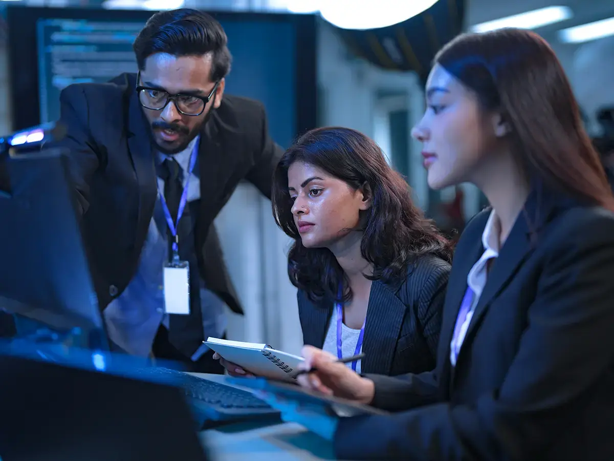 A man and two women in business attire work intently at a computer in an office