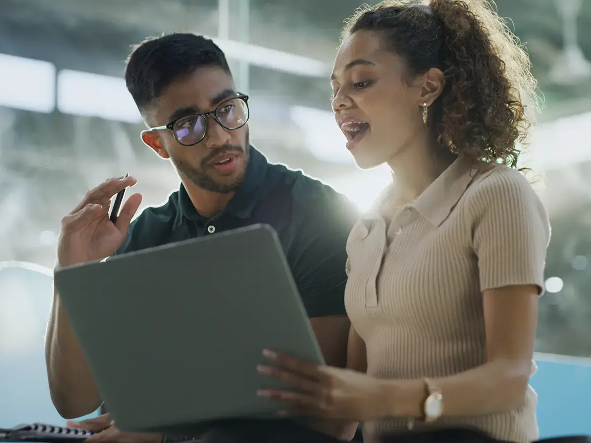 A man and woman are sitting together, engaged in a friendly conversation while looking at a laptop screen