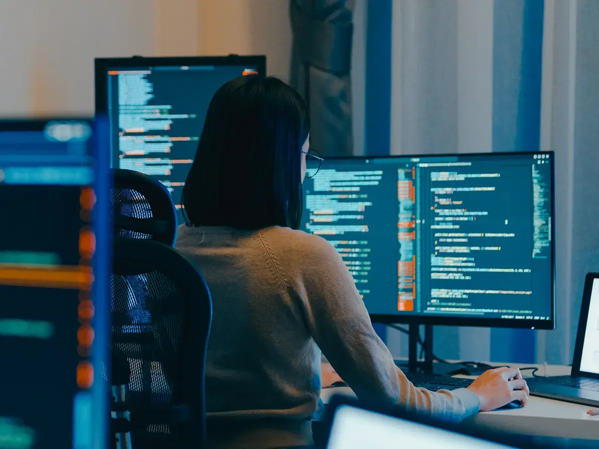 A person seated at a desk surrounded by multiple monitors displaying lines of code