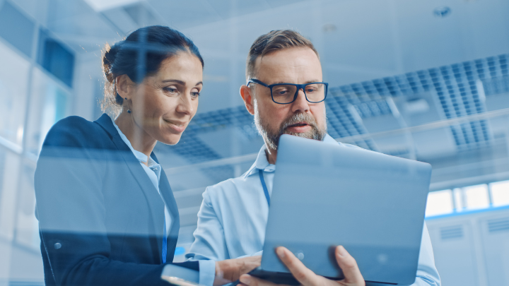 A woman and a man work together in an office, collaborating on a project while looking at the same laptop screen
