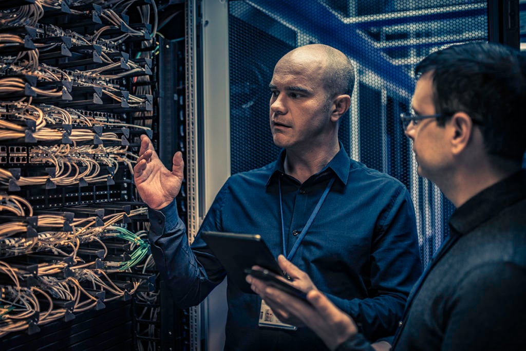 Two IT professionals stand in a server room; one holds a tablet while the other gestures as they discuss network cabling