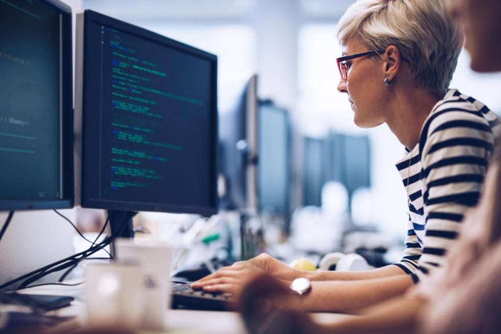 A woman with short hair, wearing a striped shirt, focuses on her work at a desk with multiple computer monitors
