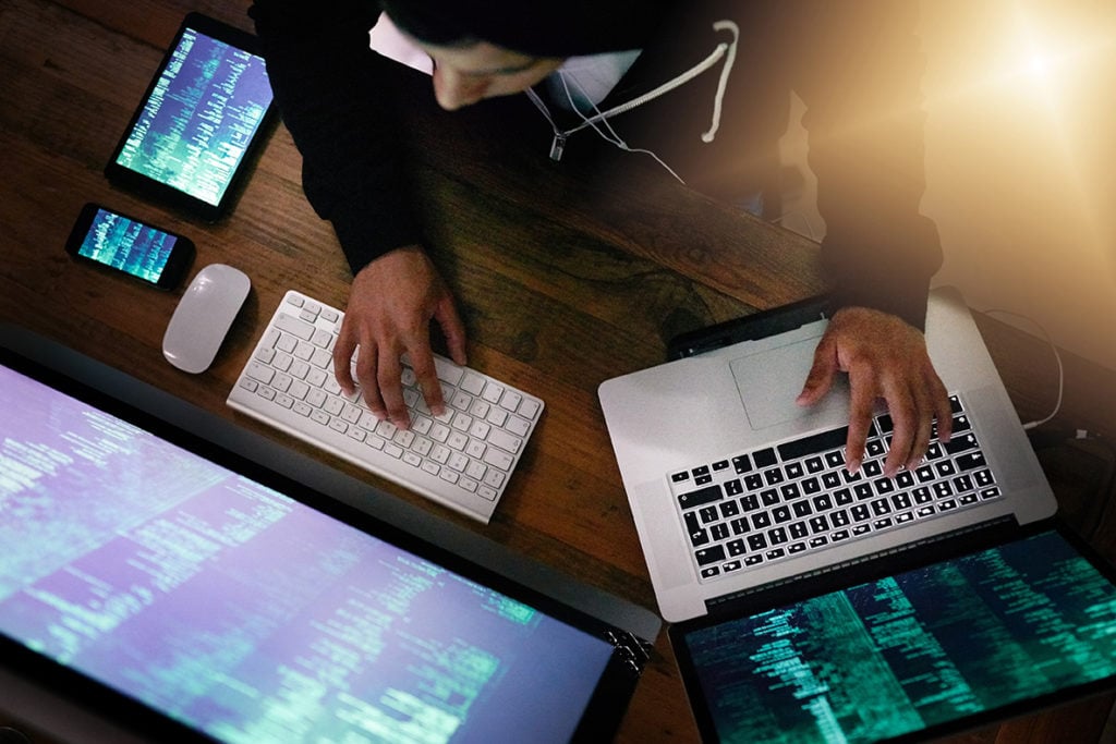 A top-down view of a person's hands typing on a multi-monitor desk setup, including a laptop and a desktop computer